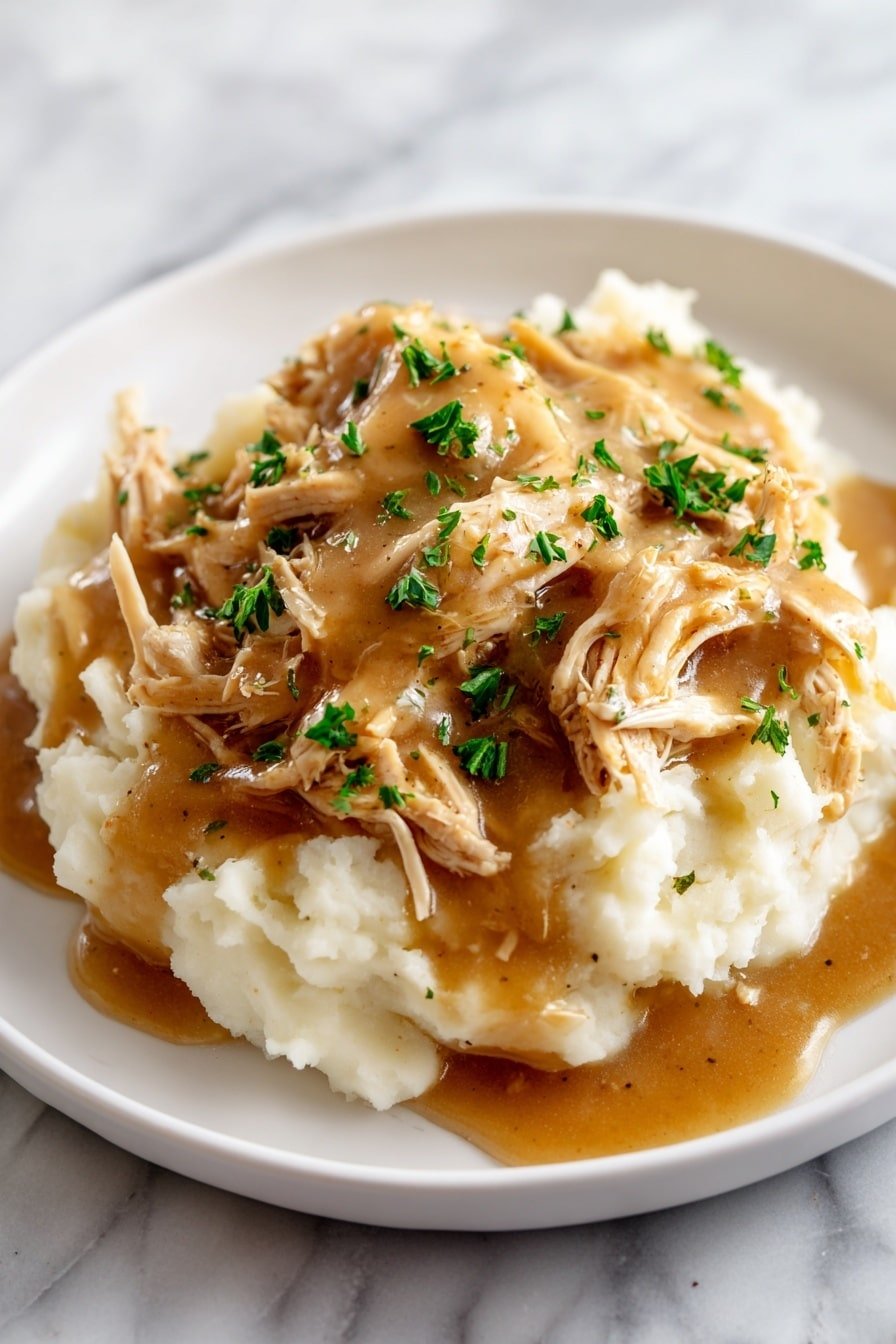 A white plate holds a serving of creamy mashed potatoes as the base layer, soft and smooth with a light off-white color. On top of the mashed potatoes, there is a layer of shredded cooked chicken, light brown with some darker brown seasoning spots, giving it a tender and juicy look. The whole dish is covered with a thick brown gravy that glistens slightly, pooling around the edges of the mashed potatoes. Small green parsley pieces are sprinkled over the chicken and gravy, adding bright color and freshness. The plate is set on a white marbled surface. Photo taken with an iphone --ar 2:3 --v 7 — Crockpot Chicken with Creamy Gravy, slow cooker chicken recipes, easy chicken dinner ideas, hearty crockpot chicken recipes, comfort food chicken recipes