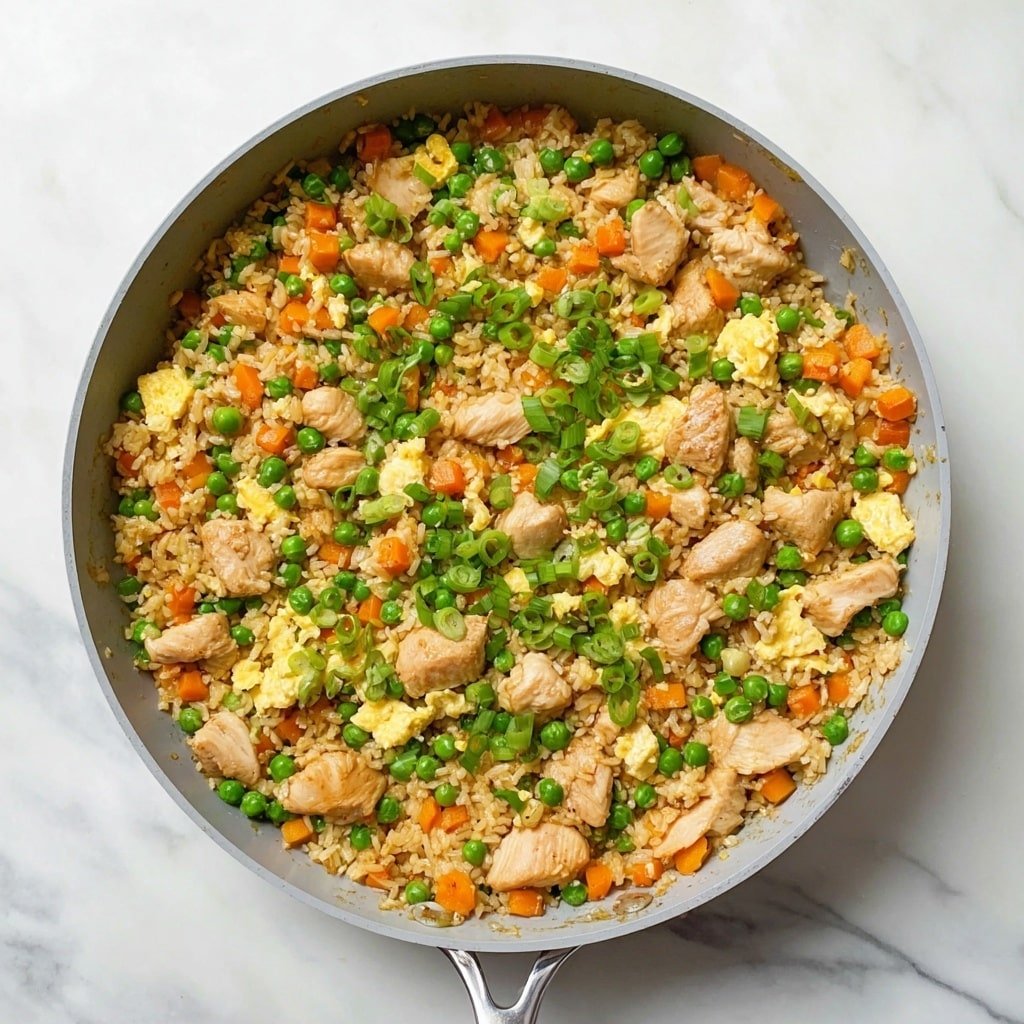 The image shows a pan filled with a colorful fried rice dish on a white marbled surface. The dish has four main layers: fluffy light brown rice spread evenly as the base, bright green peas scattered throughout, small orange carrot cubes spread evenly, and light yellow scrambled eggs mixed in. Medium-sized chunks of cooked chicken with a light golden color are evenly distributed across the pan. The dish is topped with chopped green onion slices adding fresh green shades on top. The pan has a light gray interior and a silver handle. Photo taken with an iphone --ar 1:1 --v 7 — Bang Bang Chicken Fried Rice, flavorful chicken fried rice recipe, quick Chinese fried rice, savory chicken and rice dish, easy weeknight fried rice
