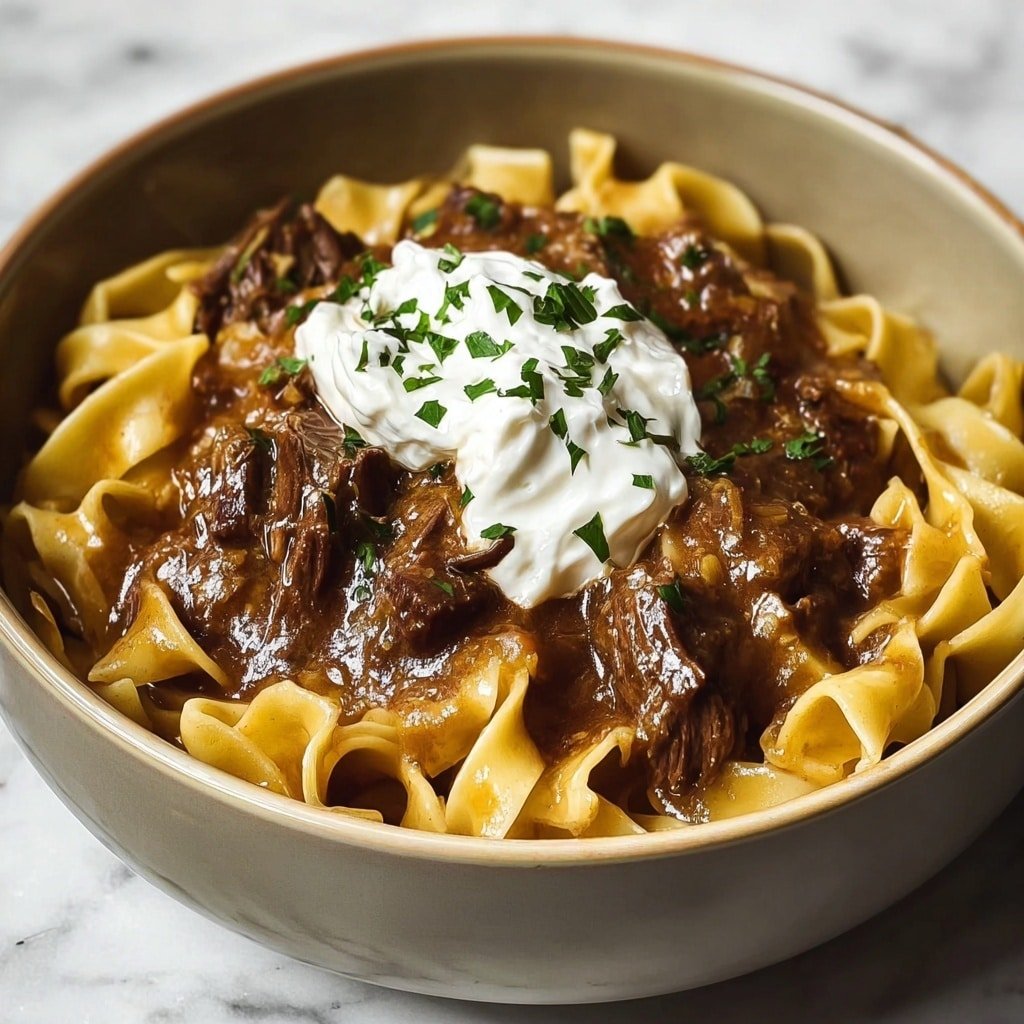 A bowl filled with wide, flat egg noodles that are a light yellow color and have a smooth texture. On top of the noodles are pieces of dark brown cooked beef in a thick, glossy brown sauce. At the center of the dish is a dollop of white creamy topping, garnished with small green herb flakes. The bowl is white and sits on a white marbled surface. photo taken with an iphone --ar 1:1 --v 7 — French Onion Beef and Noodles Slow Cooker, easy slow cooker beef recipes, comfort food recipes with beef, slow cooker beef and onion dishes, hearty weeknight dinners